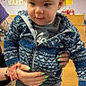 Zander is registered to the contest to win money with this photo: adult_hands, birthday_sign, blue, casual_clothing, child, curious_expression, face, green_nail_polish, head, holding, indoor, jacket, long_sleeve, pants, patterned_clothing, person, playroom, toddler, wooden_floor, young_child