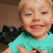 Cameron is registered to the contest to win money with this photo: child, smiling, blonde_hair, blue_eyes, tshirt, hands, indoor, living_room, couch, toy, happy, portrait, person, closeup, cute, playful, young_child, casual_clothing, home, skin