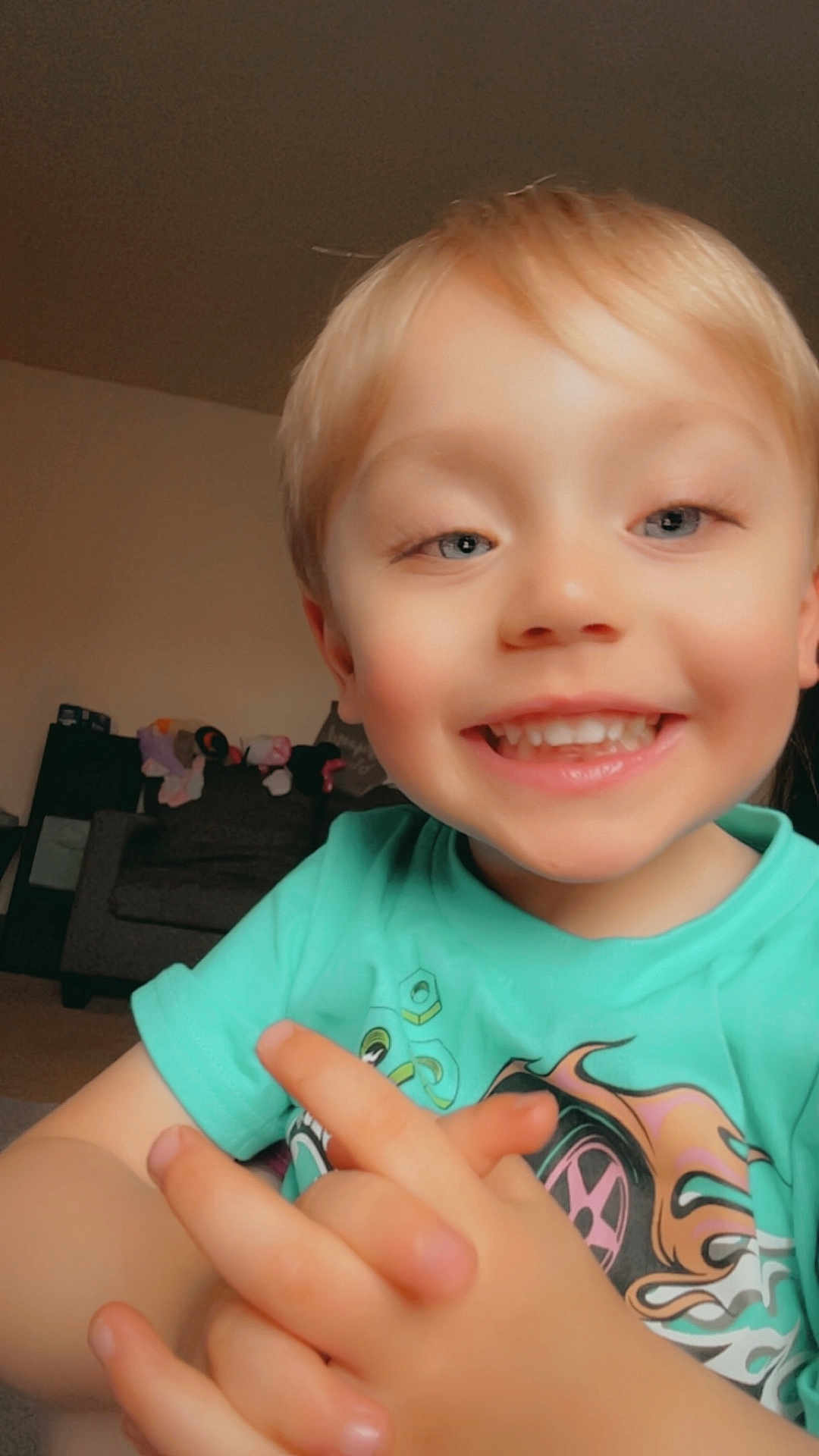 Cameron is registered to the contest to win money with this photo: child, smiling, blonde_hair, blue_eyes, tshirt, hands, indoor, living_room, couch, toy, happy, portrait, person, closeup, cute, playful, young_child, casual_clothing, home, skin