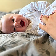 Cohen is registered to the contest to win money with this photo: baby, infant, smile, laughing, hand, holding, blanket, bed, white_clothing, face, skin, happy, closeup, person, child, indoors, human_hand, joy, cute, playful