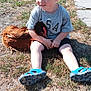 child, toddler, curly_hair, blond_hair, gray_tshirt, blue_sandals, chicken, animal, grass, outdoor, sunlight, vehicle_tire, nature, sitting, hugging, pet, rural, summer, casual_clothing, young_child