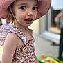 child, toddler, pink_hat, floral_dress, outdoor, portrait, curious_expression, summer, sunhat, face, eye_contact, closeup, soft_background, person, smile, cute, young_child, daylight, fashion, playful