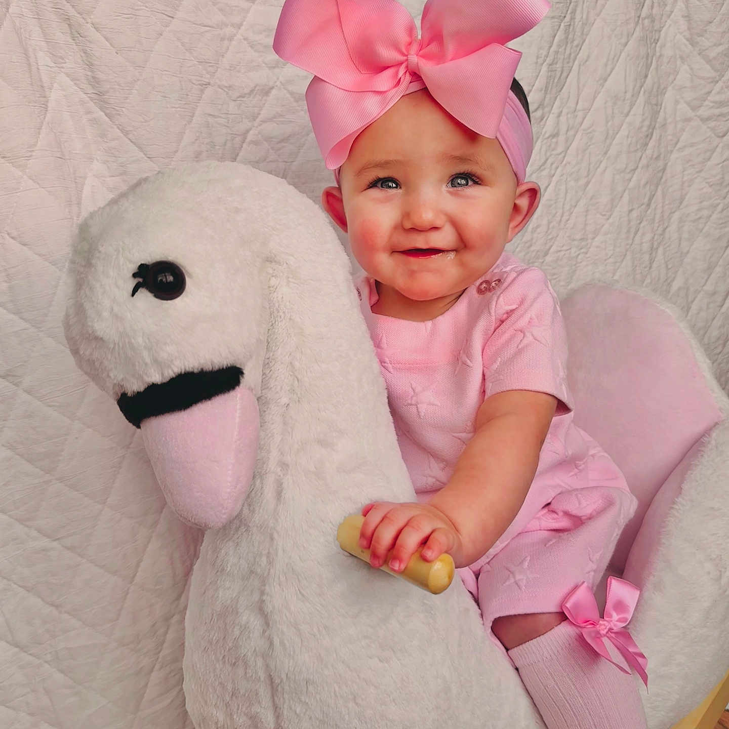 Lunden is registered to the contest to win money with this photo: baby, child, child_sitting, cute, face, floor, furniture, hand, happy, headband, indoor, pink_bow, pink_clothing, plush_toy, portrait, quilted_background, smiling, sock, swan_rocker, toy
