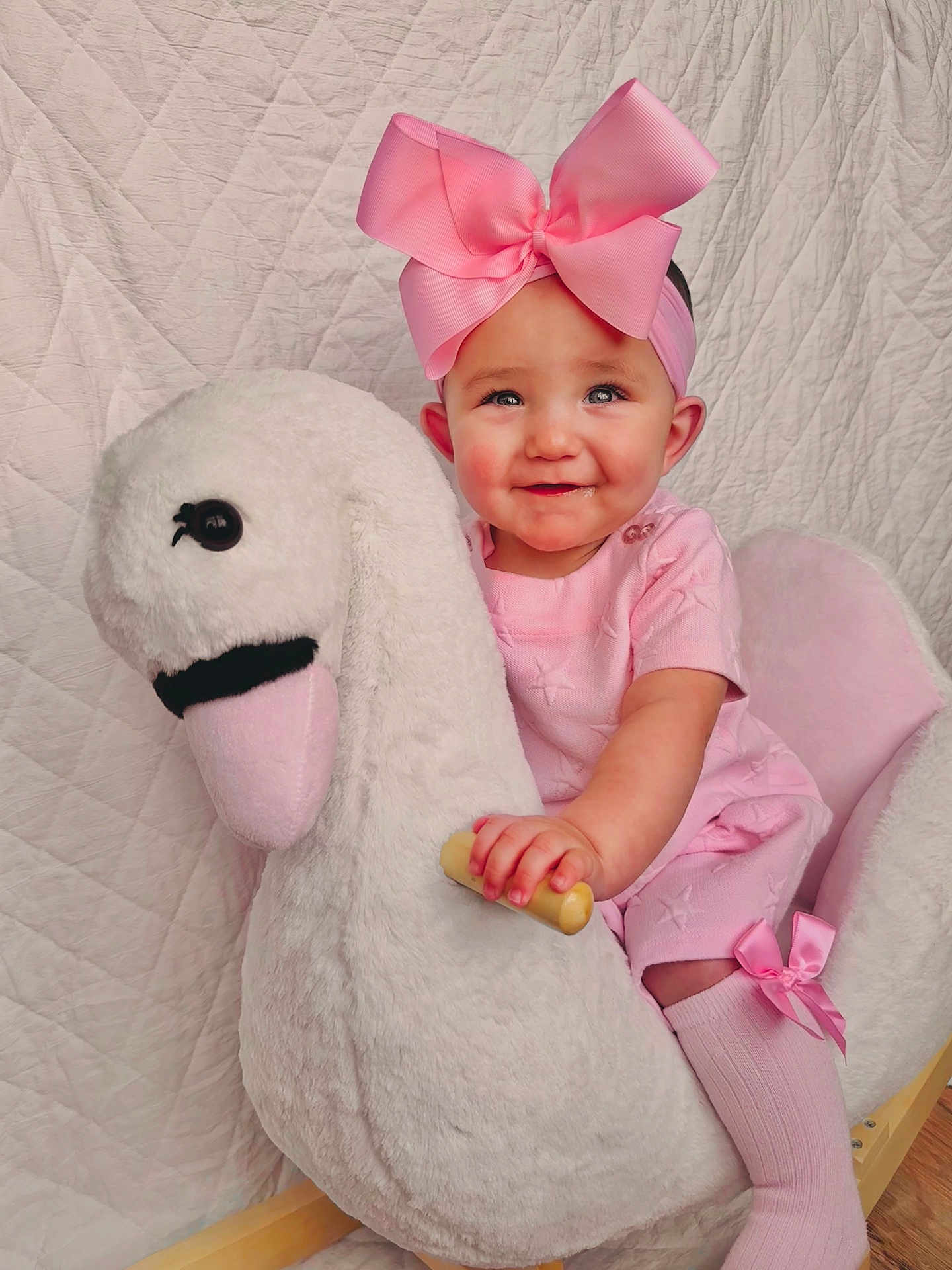 Lunden is registered to the contest to win money with this photo: baby, child, pink_bow, pink_clothing, swan_rocker, toy, smiling, cute, indoor, child_sitting, headband, sock, plush_toy, furniture, portrait, happy, face, hand, floor, quilted_background