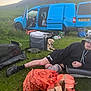 Harley is registered to the contest to win money with this photo: man, dog, golden_retriever, van, camping, grass, firewood, outdoor, tent, crate, hill, backpack, casual_clothing, relaxing, nature, pet, vehicle, leisure, daylight, hat
