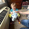 air_conditioner, carpet, child, clothing, colorful_toy, dresser, elephant, hand, happy, home, indoor, jeans, laundry_basket, person, plush_toy, ride_on_toy, smiling, toddler, toy, window