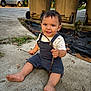 toddler, child, barefoot, stick, sidewalk, trash_bins, outdoor, casual_clothing, happy, smiling, sitting, concrete, vehicle, daylight, nature, plants, playful, young_child, person, curly_hair