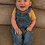 baby, smiling, denim_overalls, yellow_shirt, couch, happy, infant, child, sitting, barefoot, cute, portrait, hair_tuft, indoor, young_child, face, hands, feet, clothing, person