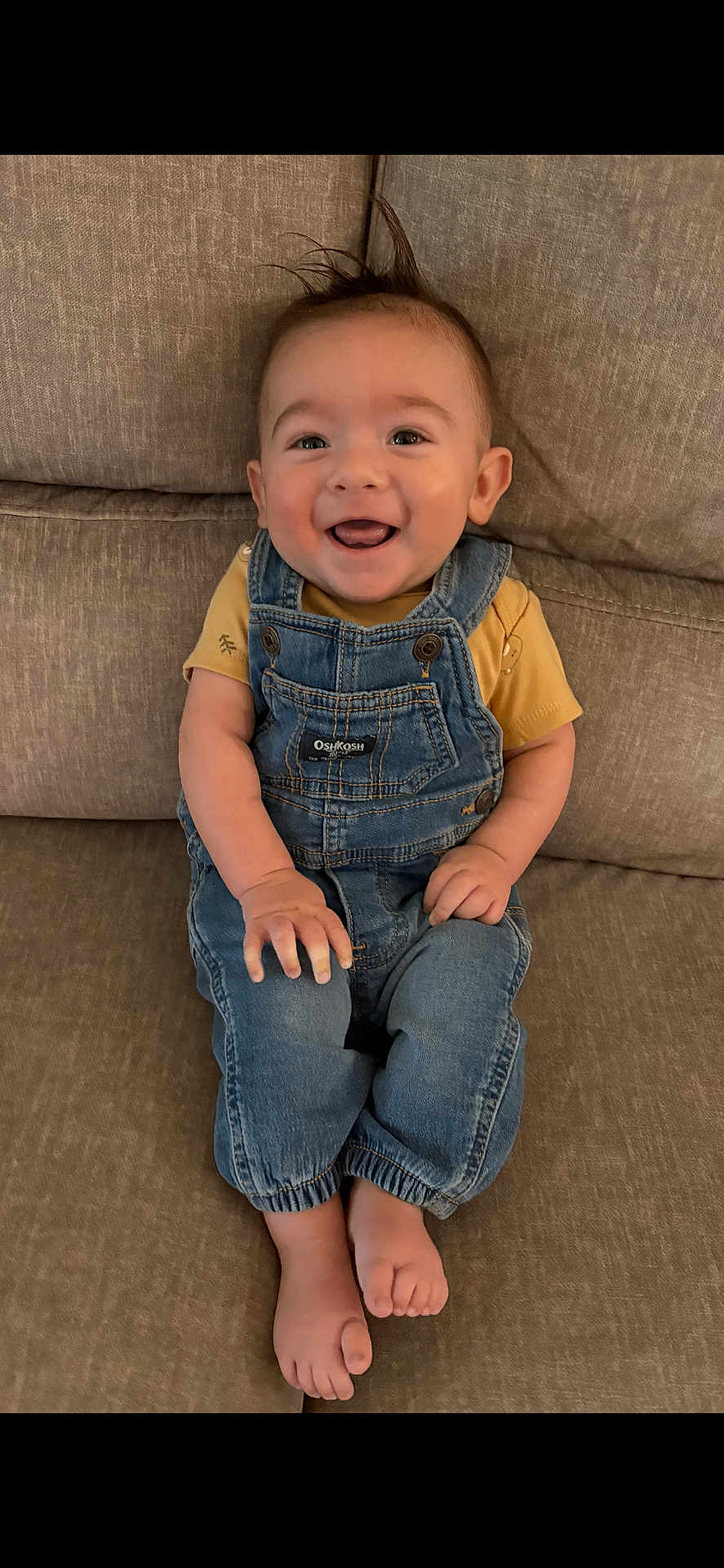 Antonio is registered to the contest to win money with this photo: baby, smiling, denim_overalls, yellow_shirt, couch, happy, infant, child, sitting, barefoot, cute, portrait, hair_tuft, indoor, young_child, face, hands, feet, clothing, person