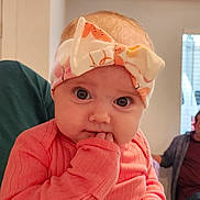 Stella is registered to the contest to win money with this photo: adult_in_background, baby, bow, child, closeup, curious, cute, expression, eyes, face, fingers, hand, headband, indoor, infant, person, pink_clothing, portrait, sitting, soft_light