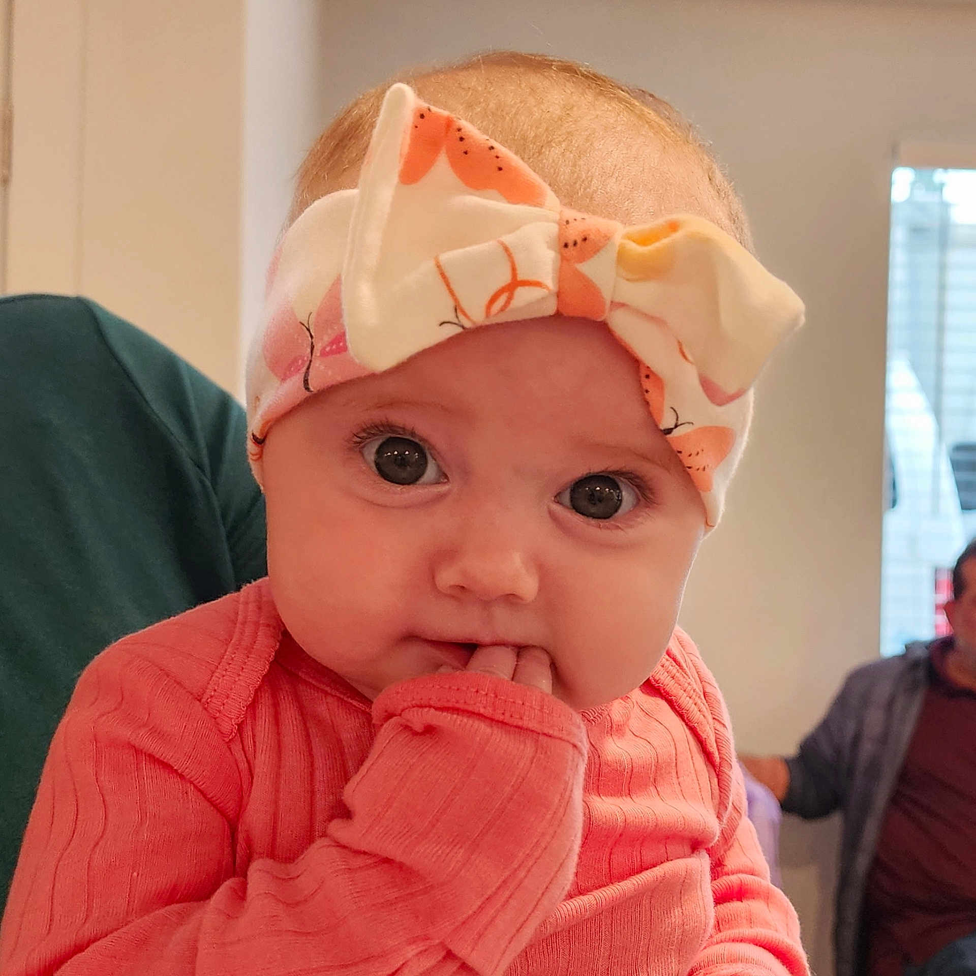 Stella is registered to the contest to win money with this photo: adult_in_background, baby, bow, child, closeup, curious, cute, expression, eyes, face, fingers, hand, headband, indoor, infant, person, pink_clothing, portrait, sitting, soft_light