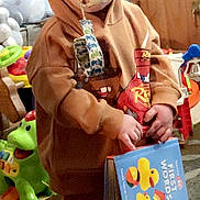 Deca is registered to the contest to win money with this photo: toddler, child, pacifier, hoodie, book, toy, playroom, dinosaur_toy, stuffed_animal, indoor, person, brown_clothing, reading, floor, carpet, cute, young_child, baby, holding, colorful
