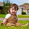 toddler, child, grass, outdoor, smiling, barefoot, overalls, short_sleeves, blue_eyes, brown_hair, house, suburban, daylight, greenery, happy, sitting, portrait, nature, casual_clothing, playful