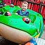 amusement_ride, background, casual_clothing, child, daylight, frog, fun, glasses, green, grip, happy, outdoor, people, play, playground, red_fence, seat, smiling, toy, young_boy