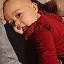 baby, infant, child, face, skin, blue_eyes, red_clothing, cushion, blanket, soft, indoors, portrait, smiling, lying_down, head, cheeks, hand, cozy, warm_colors, playful