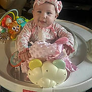 Trinity is registered to the contest to win money with this photo: baby, infant, child, smiling, headband, pink_clothing, plush_toy, baby_activity_center, toys, indoor, living_room, seat, cute, happy, playtime, person, face, closeup, childhood, soft_toy