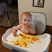 Ellie is registered to the contest to win money with this photo: baby, child, highchair, tray, food, macaroni, peas, mess, eating, socks, bare_torso, face, big_eyes, wooden_floor, framed_photos, wall, living_room, curtain, baby_gate, stool