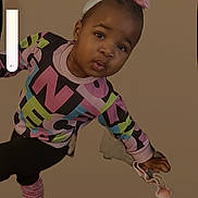 Lariah is registered to the contest to win money with this photo: child, toddler, portrait, indoor, standing, leaning, pouting, face, smiling, hair_bow, headband, colorful_clothing, patterned_shirt, pants, striped_socks, sneakers, shoe, hand, phone_screenshot, volume_indicator