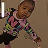 child, toddler, portrait, indoor, standing, leaning, pouting, face, smiling, hair_bow, headband, colorful_clothing, patterned_shirt, pants, striped_socks, sneakers, shoe, hand, phone_screenshot, volume_indicator