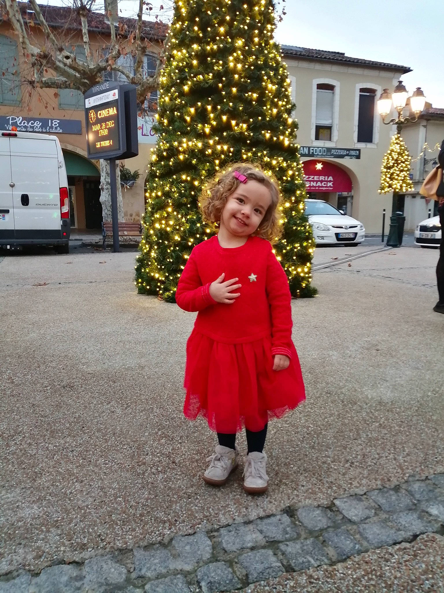 Mélina participe au concours pour gagner de l'argent avec cette photo : christmas_ornament, christmas_tree, city, event, flooring, fun, holiday, human_settlement, joy, leaf, leisure, light_commercial_vehicle, pedestrian, person, plant, road, road_surface, standing, street, toddler