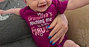 Luna joined the competition — help win amazing prizes! baby, child, smile, purple_onesie, pink_headband, adult_hand, blue_nails, couch, indoor, happy, person, sitting, furniture, reflection, cabinet, skin, legs, text_on_clothing, casual, home