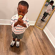 Saint is registered to the contest to win money with this photo: child, boy, toddler, standing, popsicle, snack, eating, striped_shirt, collared_shirt, shorts, sneakers, mirror, reflection, indoor, hardwood_floor, home, portrait, fashion, looking_down, smiling