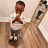 child, boy, toddler, standing, popsicle, snack, eating, striped_shirt, collared_shirt, shorts, sneakers, mirror, reflection, indoor, hardwood_floor, home, portrait, fashion, looking_down, smiling