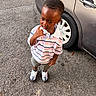 Saint is registered to the contest to win money with this photo: child, toddler, boy, portrait, outdoor, standing, car, parked_car, driveway, asphalt, striped_shirt, polo_shirt, shorts, sneakers, fashion, thoughtful, hand_on_chin, dark_skin, automobile, street