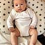 baby, infant, onesie, cushion, star_pattern, bed, curly_hair, skin, foot, hand, face, indoor, young_child, relaxed, expression, soft_lighting, technology, smartphone, white_clothing, comfort