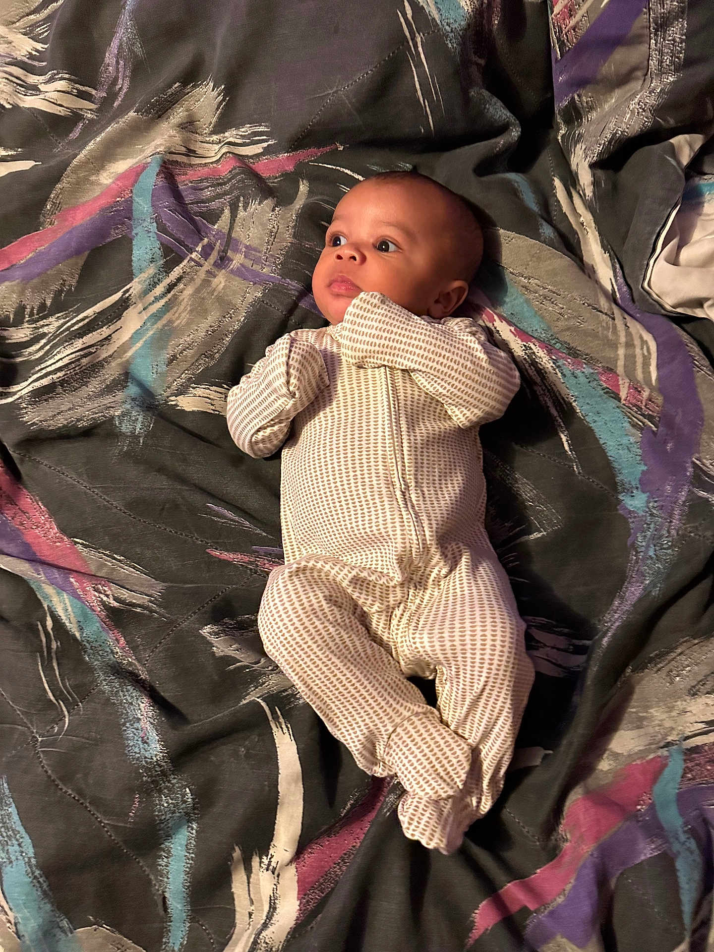Elias is registered to the contest to win money with this photo: baby, infant, onesie, blanket, pattern, laying_down, curious, face, person, child, indoor, fabric, colorful, expression, sleepwear, newborn, cute, soft, comfort, portrait