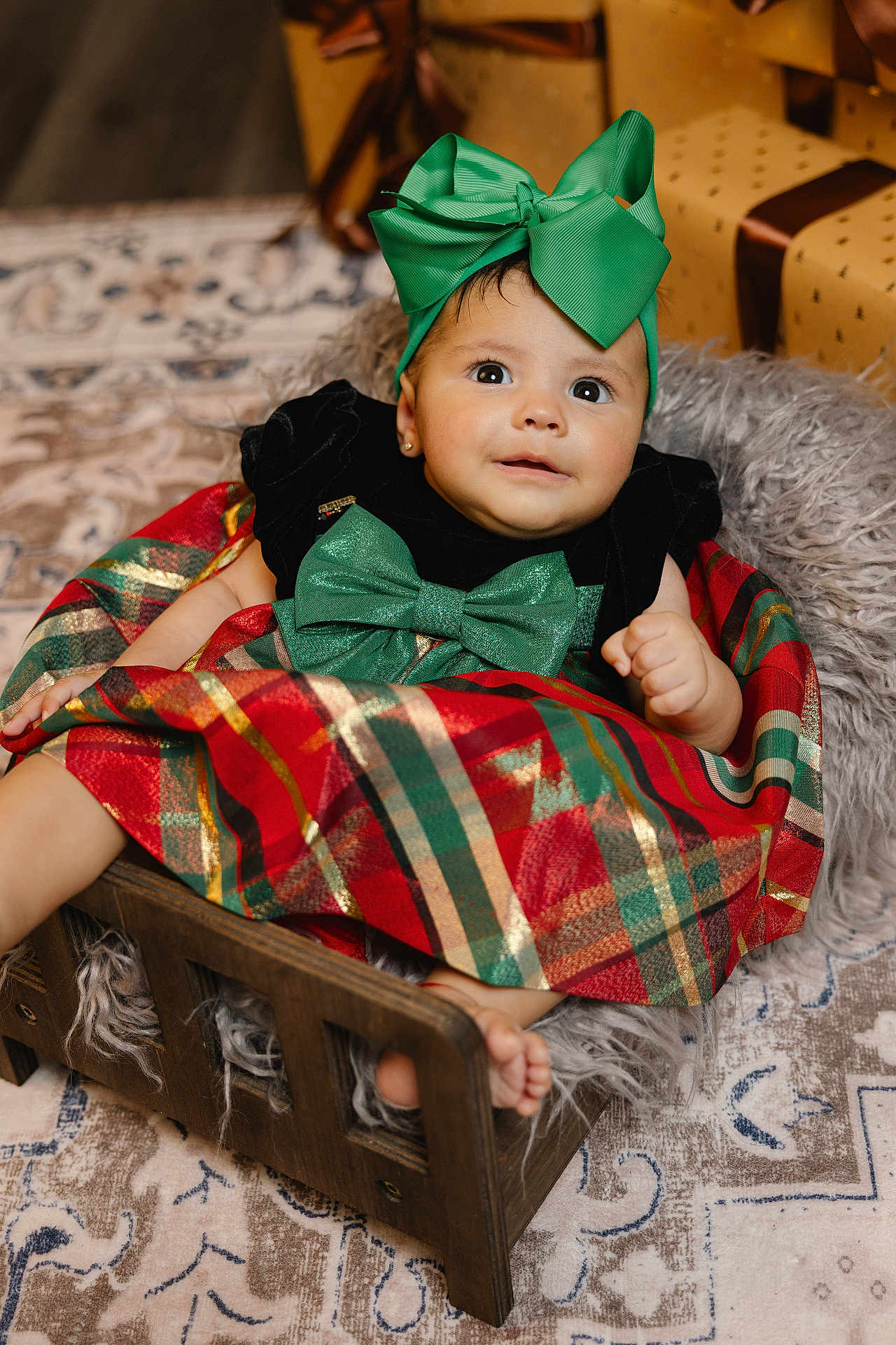 Angelina joined the competition — help win amazing prizes! baby, child, infant, green_bow, plaid_dress, wooden_bed, blanket, gift_box, earrings, holiday, festive, carpet, smiling, cute, indoors, portrait, wrapped_presents, floor_rug, cozy, happy