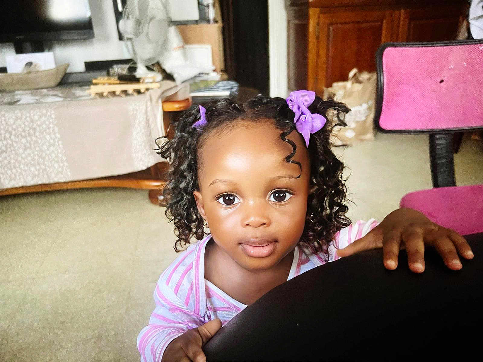 Lanaëlle a rejoint le concours — aidez-le/la à gagner de superbes lots ! child, toddler, curly_hair, purple_bows, indoor, chair, hand, big_eyes, curious, striped_clothing, home, carpet, table, fan, wooden_furniture, person, portrait, cute, face, young