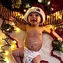 Quincey joined the competition — help win amazing prizes! baby, infant, diaper, santa_hat, christmas_lights, holiday, festive, tinsel, red_garland, smiling, happy, holiday_decor, celebration, cute, lying_down, winter, indoors, glowing_lights, joyful, festivity