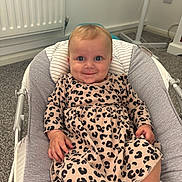 Mabel is registered to the contest to win money with this photo: baby, infant, smile, face, blue_eyes, leopard_print, dress, baby_rocker, indoor, carpet, radiator, hand, legs, cute, child, person, happy, seated, clothing, home