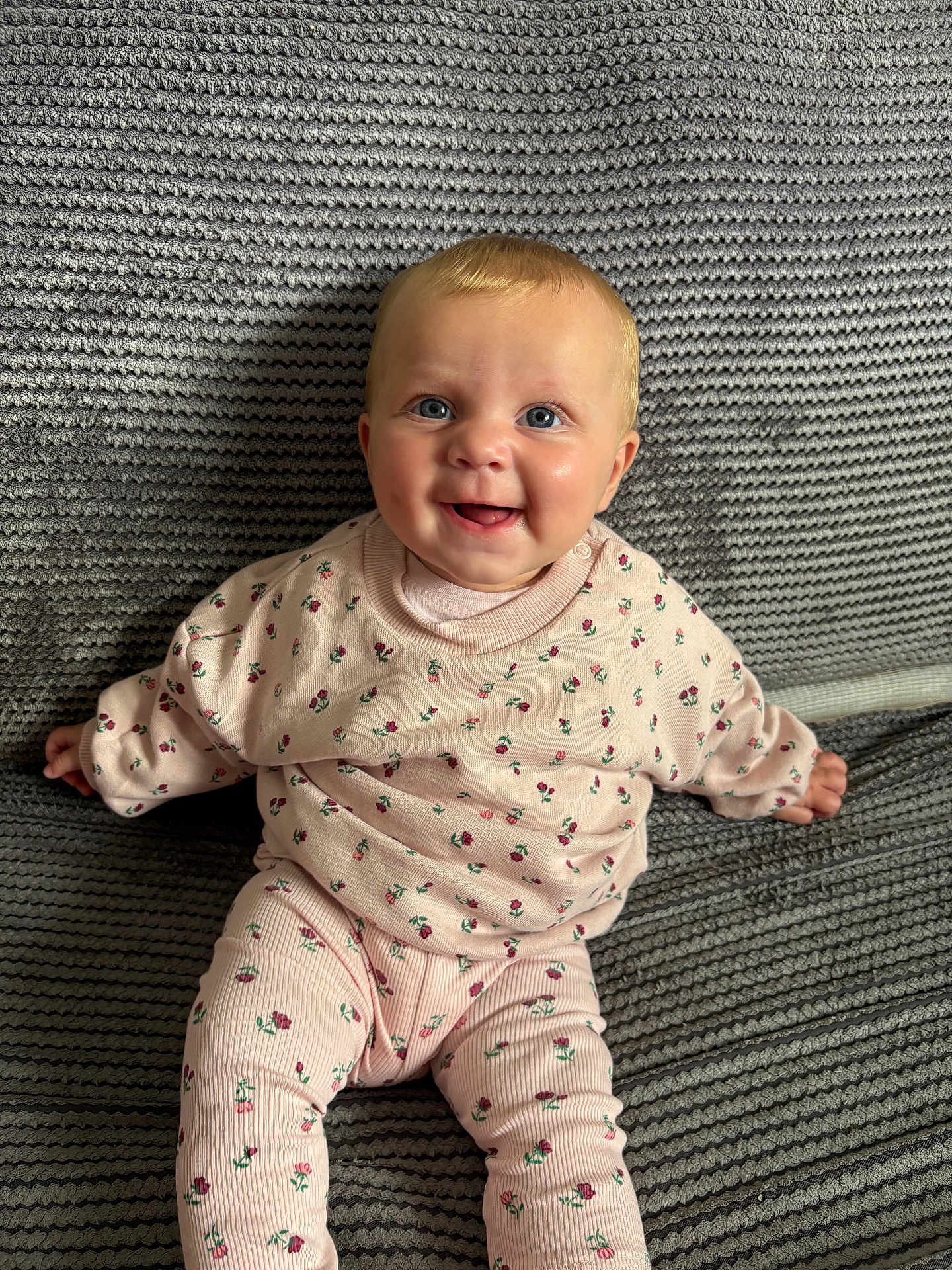 Mabel is registered to the contest to win money with this photo: baby, child, smiling, blue_eyes, pink_clothing, floral_pattern, knitted_blanket, sitting, cute, portrait, indoor, happy, infant, cozy, soft_texture, young_child, face, head, hands, legs