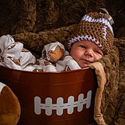 Ellianna joined the competition — help win amazing prizes! baby, infant, bucket, football, football_hat, pajamas, plush_blanket, soft_texture, cute, cozy, child, portrait, knitted_hat, sports_theme, brown, sleepy, resting, toy, indoor, cute_outfit