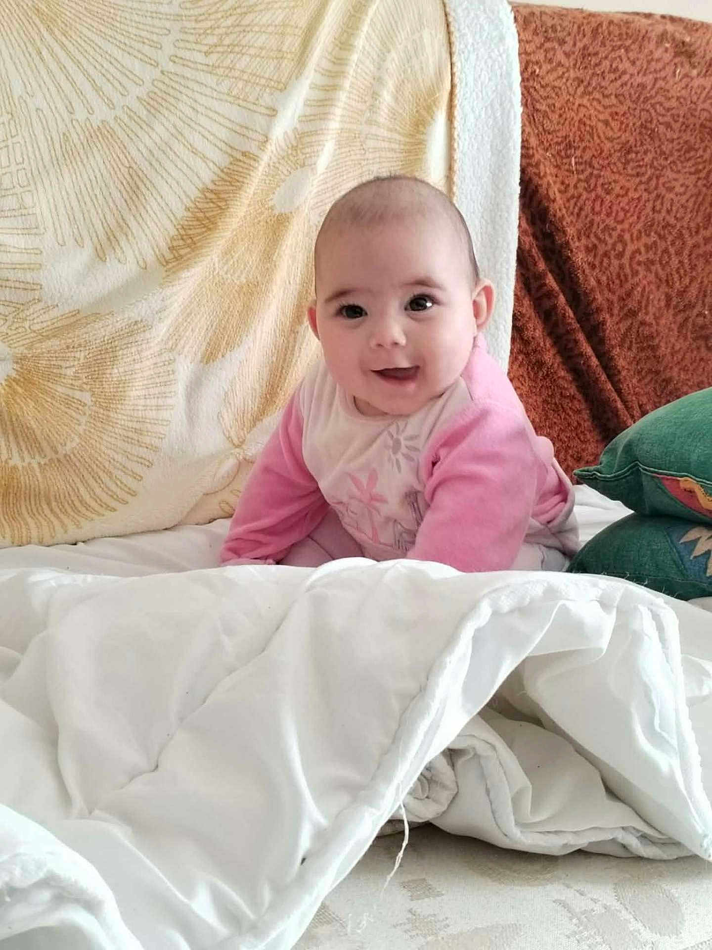 Kaïlyne a rejoint le concours — aidez-le/la à gagner de superbes lots ! baby, child, smiling, pink_clothing, white_blanket, cushion, bed, indoor, cute, portrait, face, young, infant, happy, soft, cozy, sitting, person, blanket, home