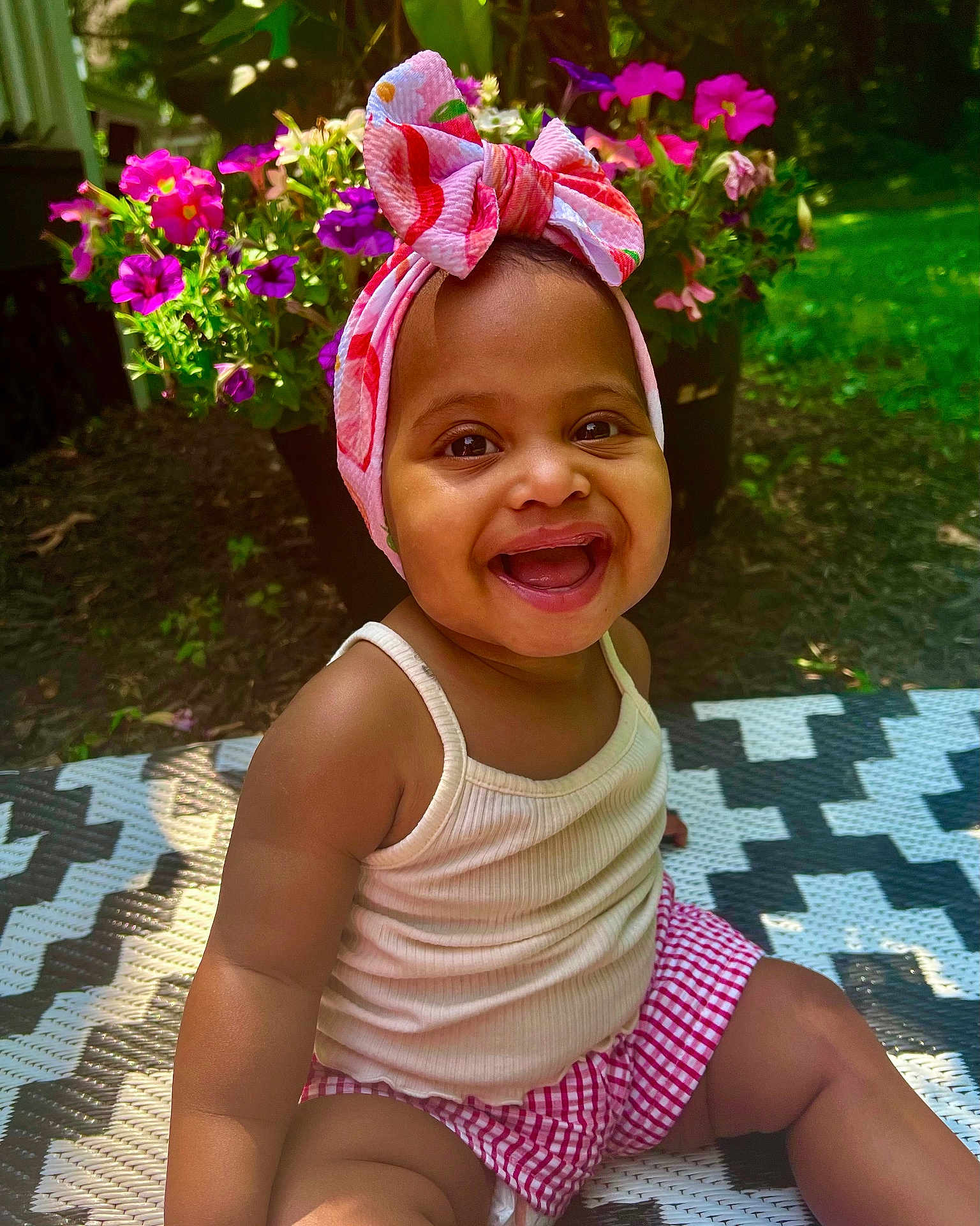 Natalia is registered to the contest to win money with this photo: baby, child, smiling, headband, outdoor, flower, greenery, patterned_mat, happy, cute, summer, sunlight, skin, tank_top, shorts, pink, red, nature, sitting, portrait