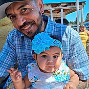 Venus is registered to the contest to win money with this photo: baby, blue_flower, building, cap, car, child, family, headband, holding, man, outdoor, parent, plaid_shirt, portrait, serious_expression, smile, sunlight, tattoo, woman, yellow_dress