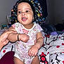 baby, child, bonnet, onesie, floral_pattern, hand, bed, blanket, smiling, happy, indoor, person, cute, infant, portrait, skin, expression, holding, bedroom, comfort