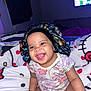 baby, smiling, sitting, bed, blanket, onesie, bonnet, face, happy, child, indoors, cute, person, furniture, television, patterned_clothing, home, colorful, portrait, cozy