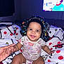 baby, bedroom, blanket, bonnet, cartoon, child, colorful, cute, face, hand, happy, holding_hands, indoor, infant, onesie, pattern, person, sitting, smiling, television