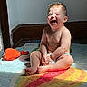 baby, child, infant, laughing, smiling, sitting, diaper, indoor, sunlight, rainbow_rug, colorful, portrait, face, barefoot, happy, playroom, floor, orange_hat, home, candid