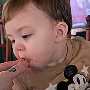 Thomas is registered to the contest to win money with this photo: baby, infant, child, face, portrait, close_up, curious, eating, finger, hand, tattoo, mickey_mouse, disney, shirt, clothing, stars, indoor, home, television, head