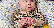 Arianna joined the competition — help win amazing prizes! baby, infant, onesie, leopard_print, pillow, floral_pattern, purple_bedding, wooden_wall, indoor, child, cute, hands, face, expression, comfort, toy, colorful, sitting, curious, portrait