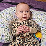 baby, infant, onesie, leopard_print, pillow, floral_pattern, purple_bedding, wooden_wall, indoor, child, cute, hands, face, expression, comfort, toy, colorful, sitting, curious, portrait