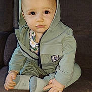 Naël participe au concours pour gagner de l'argent avec cette photo : toddler, child, hoodie, green_clothing, socks, indoor, sofa, brown_sofa, seated, baby, cute, curious, small_hands, face, portrait, young_child, casual_wear, cozy, home, expression