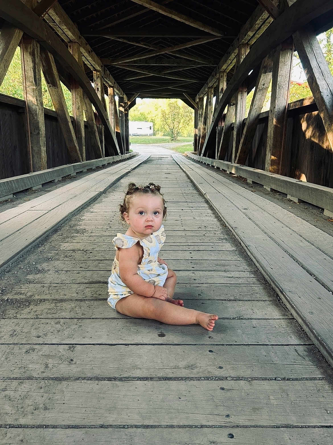 Laelynn is registered to the contest to win money with this photo: toddler, child, covered_bridge, wooden_floor, outdoor, sitting, curly_hair, pigtails, summer_clothes, innocent, portrait, nature, daylight, rustic, wood, planks, expression, looking_up, alone, quiet
