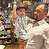 baby, man, child, hat, shirt, indoor, store, toys, smile, holding, person, wooden_floor, shelf, clothing, beard, facial_hair, light, background, happy, portrait