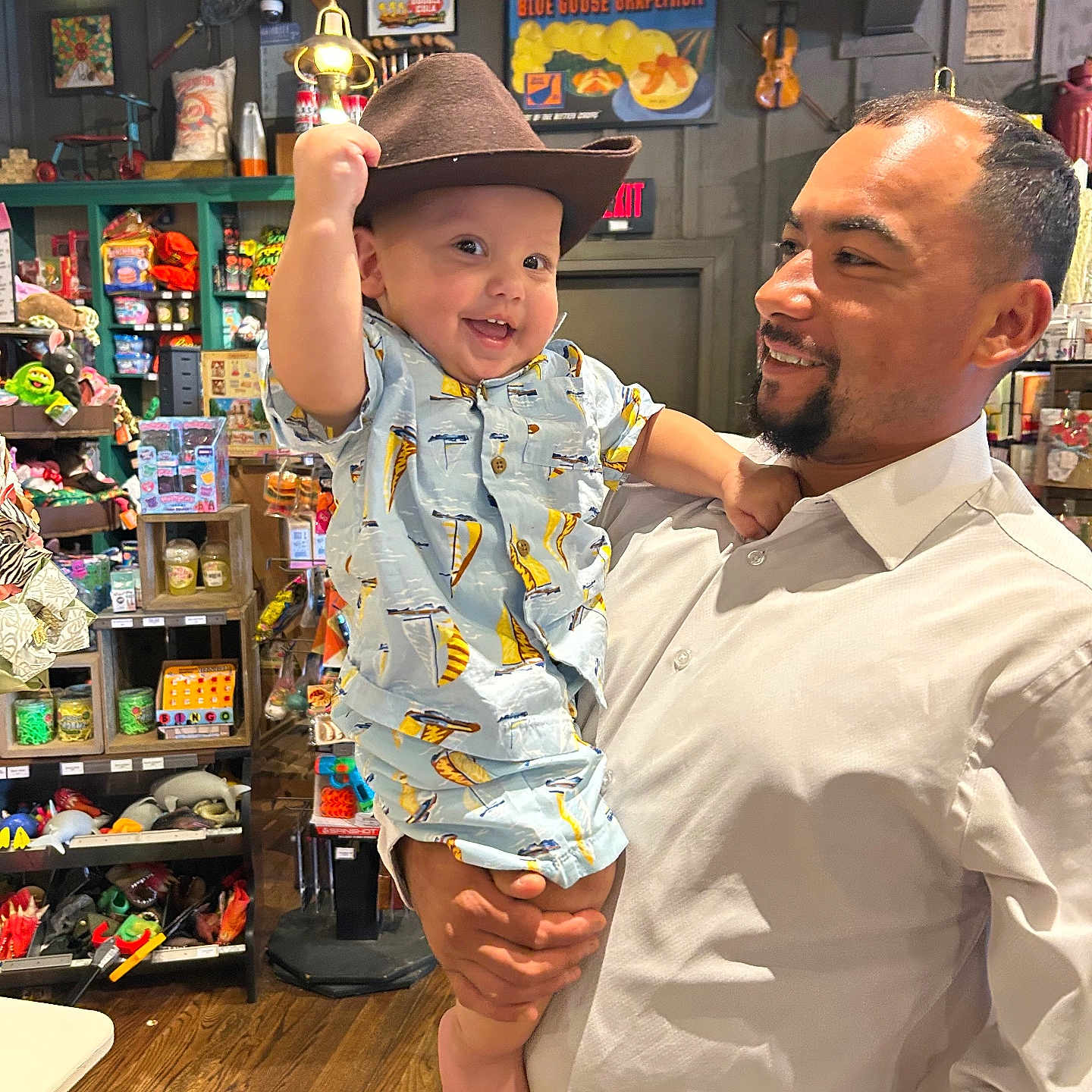 Misael is registered to the contest to win money with this photo: baby, man, child, hat, shirt, indoor, store, toys, smile, holding, person, wooden_floor, shelf, clothing, beard, facial_hair, light, background, happy, portrait